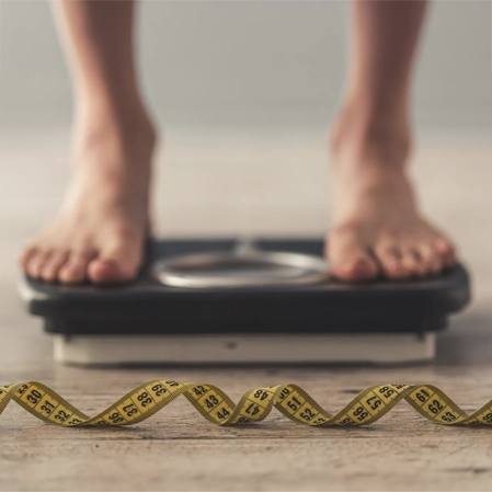 A person stands on a scale with a measuring tape in front, symbolizing weight‑management efforts.